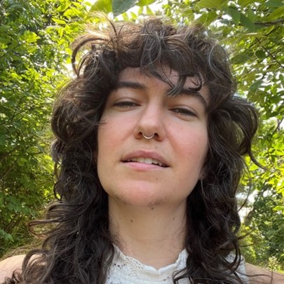 Person with curly hair and a nose ring outdoors among greenery.